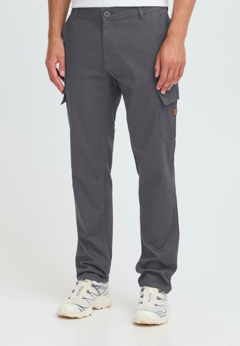 Man wearing dark gray cargo pants with flap pockets and white athletic shoes standing against a plain background.