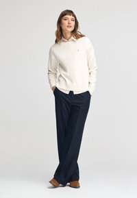 Maglione chiaro in crema con scollo tondo, indossato sopra una camicia button-up a quadri rosa, abbinato a pantaloni larghi blu navy e scarpe in suede marroni.