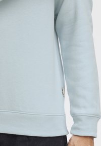 Felpa azzurra chiara con una texture morbida, polsini e orlo a costine, con una piccola etichetta logata sul lato. Primo piano del manica e dell'orlo.