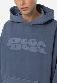 Hanorac cu glugă de culoare albastră, fabricat dintr-un material moale, având un logo texturat proeminent de culoare albastră deschisă pe piept și o buzunar tip kangaroo.