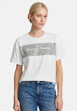Mujer rubia con una camiseta blanca de Maison Caelle Paris con un diseño rectangular plateado y brillante, combinada con vaqueros azules de talle alto.