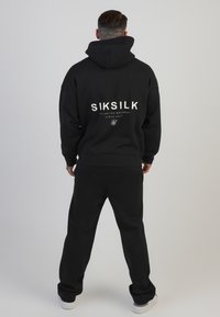 Svart hoodie och matchande byxor, med vit "SIKSILK" logotyp på ryggen, tillverkade av mjukt tyg.