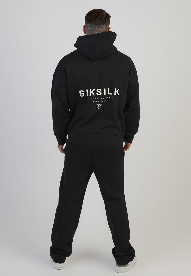 Svart hoodie och matchande byxor, med vit "SIKSILK" logotyp på ryggen, tillverkade av mjukt tyg.