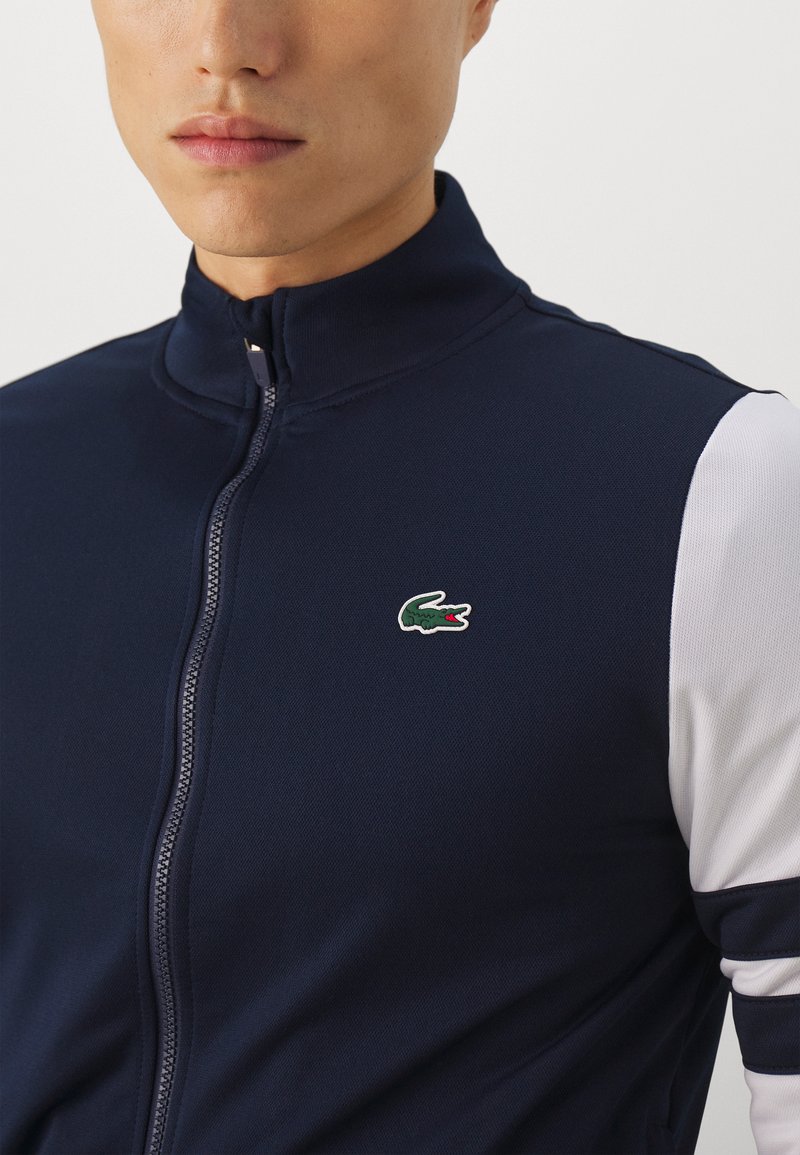 Veste zippée bleu marine avec un col haut, présentant des accents blancs sur les manches et un petit logo brodé vert sur la poitrine. Texture lisse.