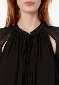 Zwarte blouse met beaded halslijn, geraffineerde stof en kwastaccent. Heeft open schouders en een lichte textuur.