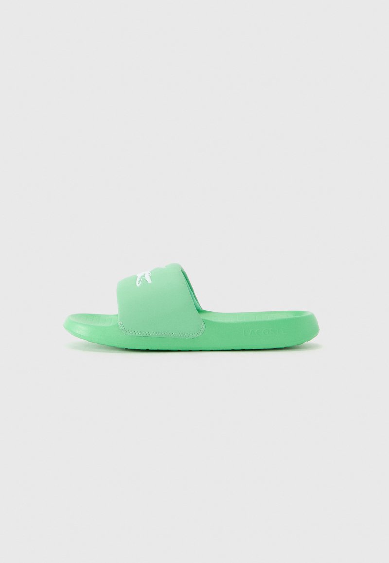 Green Lacoste slide sandal with wide strap featuring white crocodile logo, textured footbed, and embossed brand name on sole.