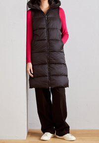 Czarna puchowa kamizelka z kapturem, zapinana na zamek z przodu, z pikowaną teksturą. Noszona na długim czerwonym topie z długim rękawem, w połączeniu z czarnymi spodniami.