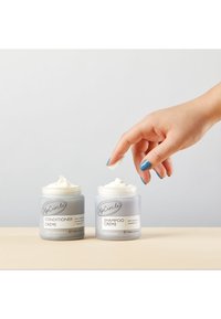 Dwa matowe szklane słoiki oznaczone jako "Conditioner Crème" i "Shampoo Crème" z białą kremową konsystencją, obok ręka sięgająca po słoik z szamponem.