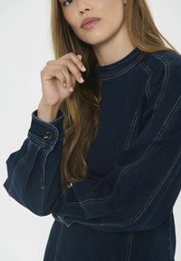 Sötétkék denimruha magas nyakkal és puffos hosszú ujjakkal. Kontrasztos varrás és gombdíszítések találhatók az ujjakon.