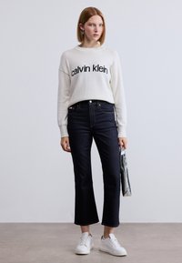 Λευκό πλεκτό πουλόβερ με το λογότυπο «calvin klein», σκούρα μπλε τζιν φαρδιάς γραμμής, λευκά αθλητικά παπούτσια, κρατώντας μεταλλικό τσαντάκι, μινιμαλιστικός σχεδιασμός.