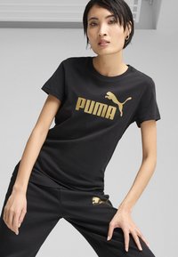 Fekete pamut póló arany "PUMA" logóval és egy ugráló pumát ábrázoló grafikával. Klasszikus kerek nyakkal és rövid ujjakkal rendelkezik.