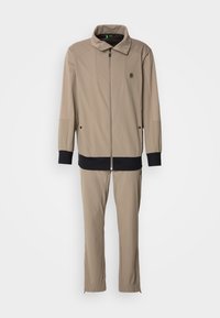 Survêtement beige deux pièces avec veste zippée munie de poignets et d'un ourlet noirs, pantalon assorti avec chevilles élastiques et logo de la marque sur la poitrine.