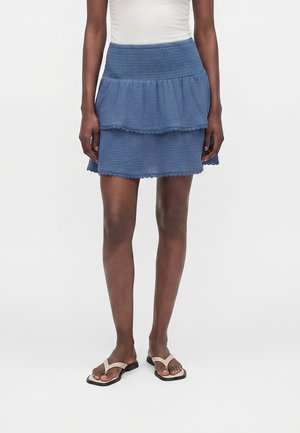 Femme portant une jupe bleue à volants avec une ceinture froncée et des sandales beige sur un fond blanc uni.