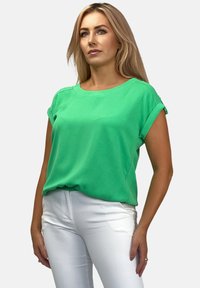 Zielona bluzka z krótkim rękawem i okrągłym dekoltem, wykonana z gładkiego materiału. Połączona z białymi dopasowanymi spodniami, prezentującymi luźny krój.