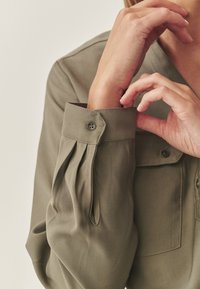 Olivegrünes Langarmshirt mit gefälteltem Bündchen, Knopfdetails und glatter Textur. Eine einzelne Brusttasche ist sichtbar.