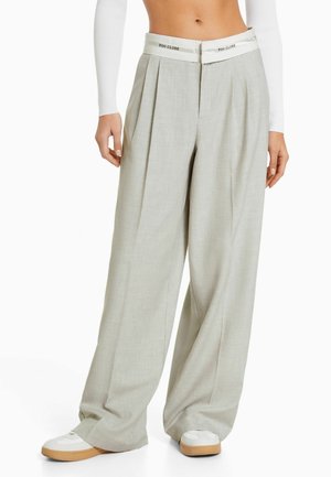 Lichtgrijze wijde broek met plooien, voorzien van een contrasterende witte tailleband met het opschrift "TOO CLOSE" en gedragen met witte sneakers.