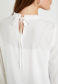 Vit blus med en texturerad volangkrage och en nyckelhålsrygg, knuten med två tygband vid halsen. Slätt, lätt material.