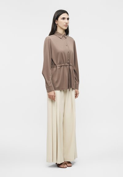 Γυναίκα που στέκεται, φορώντας μια μακρυμάνικη μπλούζα σε αποχρώσεις του ταούπε με κουμπιά και ζώνη στη μέση, συνδυασμένη με κρεμ παντελόνες φαρδιάς γραμμής και καφέ σανδάλια με ανοιχτά δάχτυλα.