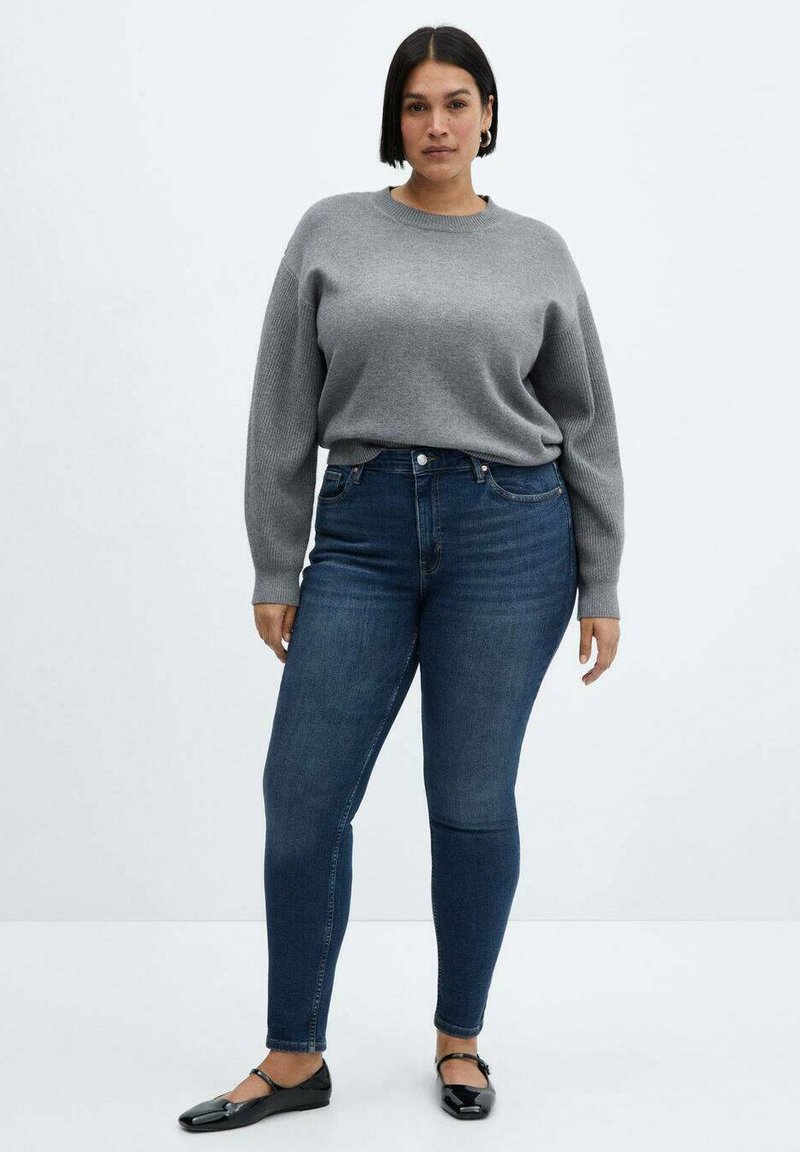 Grauer Pullover mit gerippter Textur, kombiniert mit dunkelblauen Skinny-Jeans. Schwarze Ballerinas werden an den Füßen getragen, vor einem schlichten Hintergrund.