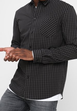 Mann trägt ein schwarz-grau kariertes Hemd mit Knöpfen und Brusttasche, darüber ein weißes Unterhemd, kombiniert mit grauen Jeans.