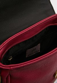 Interno della borsa in pelle sintetica bordeaux con fodera nera, dotato di una tasca con zip e un'etichetta con la scritta "ANNA FIELD." Superficie esterna testurizzata.