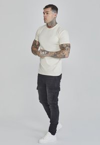 Tricou bej ajustat, pantaloni cargo negri cu buzunare laterale și adidași albi. Modelul are tatuaje, stând în fața unui fundal deschis.