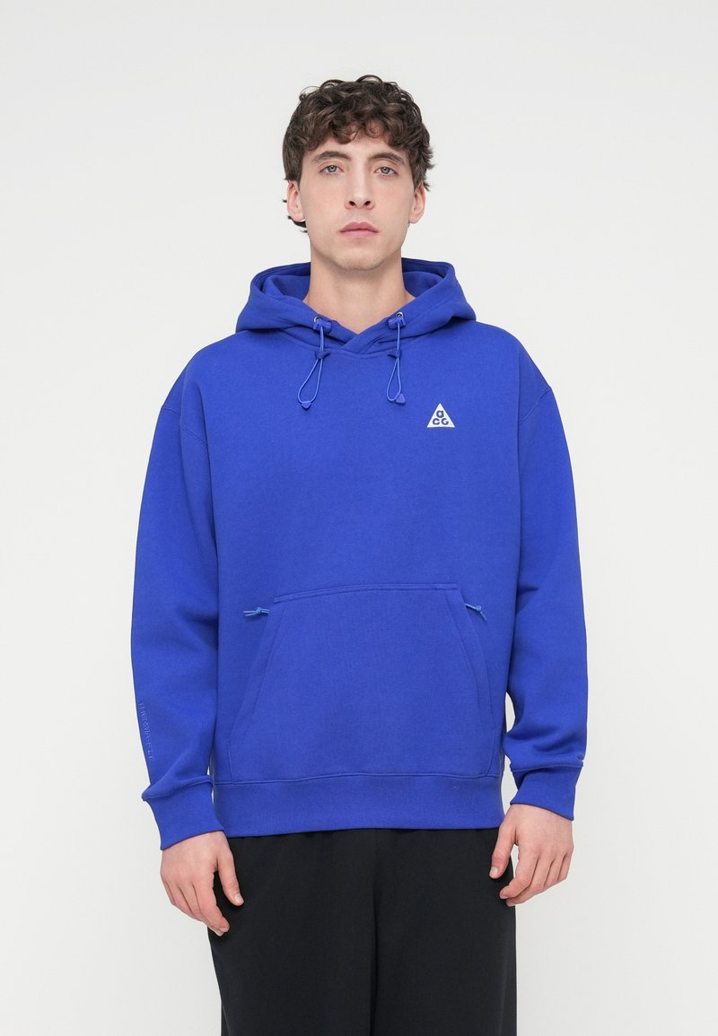 Μπλε φούτερ με κουκούλα και κορδόνια, τσέπη καγκουρό και μικρό λευκό λογότυπο. Κατασκευασμένο από μαλακό ύφασμα, διαθέτει ριμπ μανσέτες και τελείωμα.