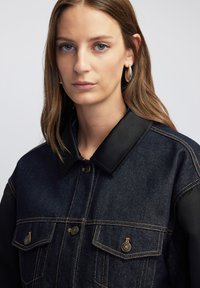 Giacca in denim blu scuro con maniche in tessuto nero, colletto ampio, bottoni in tono dorato e cuciture a contrasto. Texture liscia e due tasche sul petto.