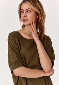 Olive zöld, rövid ujjú felső széles nyakkivágással és puha, könnyű anyagból. Az ujjak a mandzsetta körül enyhén ráncoltak.
