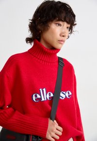 Rode gebreide trui met een hoge ribbelkraag, voorzien van blauwe en witte "ellesse" tekst op de voorkant. Model draagt een zwarte schoudertas.