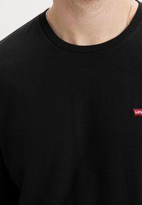 Camiseta de algodón negra con un cuello redondo y mangas cortas, que presenta un pequeño logotipo rojo de Levi's en el área izquierda del pecho.