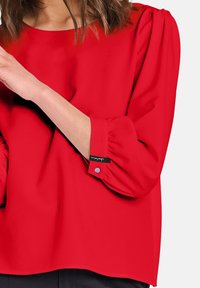 Blusa rossa con maniche a trequarti, caratterizzata da un collo rotondo e delicati dettagli sulle spalle a palloncino. Polsino con bottoni e etichetta con logo.