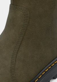 Botte en cuir vert olive avec des accents cousus, une forme arrondie et une finition texturée. La semelle présente un liseré jaune contrastant.