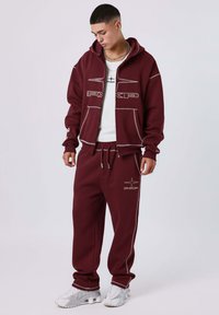 Felpa cappuccio bordeaux e pantaloni jogger abbinati, caratterizzati da cuciture bianche e dettagli del logo. Tessuto morbido e vestibilità rilassata, abbinati a sneakers bianche.