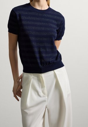 Kvinde iført en marineblå strikket top med korte ærmer og diskrete horisontale striber samt hvide højtaljede plisserede bukser mod en ensfarvet baggrund.