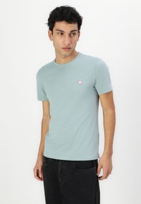 Lyseblå t-shirts med korte ærmer lavet af bomuld, der har et lille rødt trekantet logo på venstre bryst. Glat tekstur, rund halsudskæring.