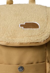 Geltona kuprinė su tekstūruotu, vilnos pavidalo atlošu, rudu logotipo lopu, metaliniu spaustuku ir lygiu audinio korpusu.