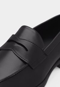 Mocassin penny en cuir noir avec finition polie, bout arrondi et une bride traversant le cou-de-pied avec un détail découpé.