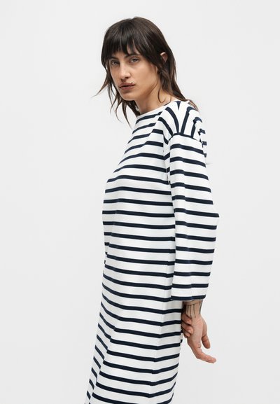 Γυναίκα με σκούρα μαλλιά που φοράει λευκό φόρεμα με οριζόντιες μαύρες ρίγες, στέκεται μπροστά σε ένα απλό ανοιχτό γκρίζο φόντο.