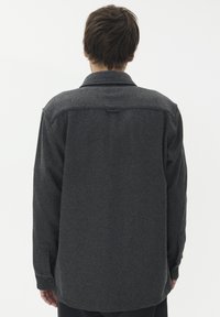 Sivi volneni srajc s dolgimi rokavi, gumbastim ovratnikom in ravnim krojem. Opoznavne lastnosti vključujejo gumbe na manšetah in šivane detajle.