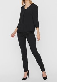 Schwarze V-Ausschnitt-Bluse mit 3/4-Ärmeln und Faltendetails, kombiniert mit schwarzen Slim-Fit-Hosen und schwarzen High Heels, vor einem neutralen Hintergrund präsentiert.