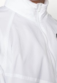 Vit lättviktsjacka med hög krage, dragkedjestängning och texturerat mönster. Har subtila detaljer och en slät yta.