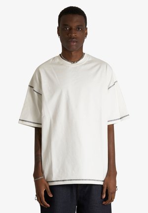 Giovane uomo con pelle scura che indossa una maglietta bianca larga con cuciture nere, pantaloni scuri, collana a catena d'argento e braccialetti, in piedi davanti a uno sfondo bianco.