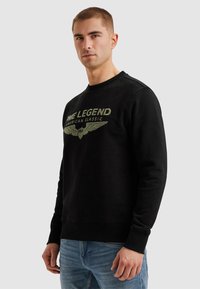 Černá mikina s výrazným grafickým potiskem v zlatém písmu a designem křídel, s žebrovaným kulatým výstřihem a manžetami, nošena s světle modrými džíny.