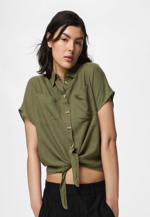Jeune femme aux cheveux foncés mi-longs portant une chemise boutonnée à manches courtes couleur vert olive, nouée à la taille, et un pantalon noir.