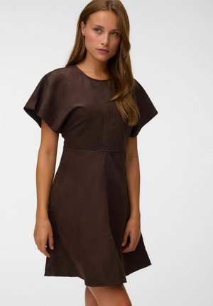 Femme aux longs cheveux bruns portant une robe marron à manches courtes, debout devant un fond clair uni.