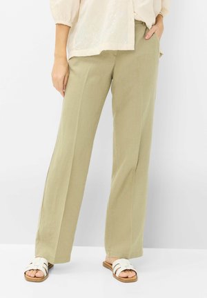 Vrouw met beige broek met wijde pijpen, witte blouse met structuur en witte open-toe platte sandalen, staand met één hand in de zak.