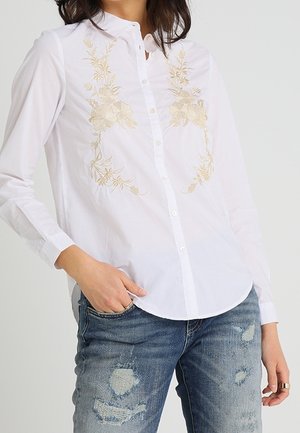 Chemise blanche boutonnée avec broderie florale beige sur la poitrine, portée avec un jean bleu délavé, main gauche du modèle dans la poche.