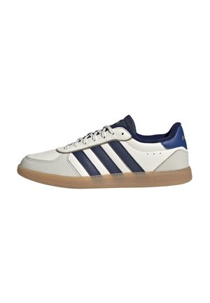 Valkoinen matala Adidas-tennari tummansinisillä raidoilla, tummansinisellä kantalapulla, kumipohjalla ja valkoisilla nauhoilla sivuprofiilissa.