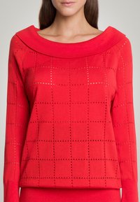 Czerwony sweter z szerokim, płaskim kołnierzem. Posiada fakturowany wzór dzianiny z kwadratowymi wycięciami w tkaninie. Długie rękawy.